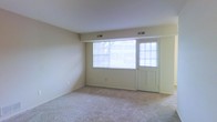 This single-floor residential space offers a clean and functional layout designed for comfortable everyday living. The main living area is a standout feature, characterized by plush wall-to-wall carpeting that adds warmth underfoot and a neutral color palette throughout, lending the space a bright and airy feel. A large picture window dominates the front wall, flanked by a paneled entry door with its own window inset, together allowing an abundance of natural light to pour into the room and offering views of the surrounding outdoor greenery. Horizontal blinds on both the window and door provide convenient light and privacy control. The smooth, light-toned walls are complemented by white trim and baseboards, contributing to the clean, understated aesthetic of the interior. Ceiling-mounted HVAC vents are positioned above the main window wall, ensuring efficient climate control throughout the space. The overall ambiance is open, calm, and move-in ready, providing a versatile blank canvas that can be easily personalized to suit a variety of interior styles and furnishing arrangements.