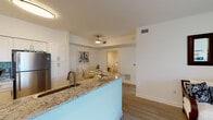 This cheerful, coastal-inspired residence is thoughtfully designed across a single floor, offering a seamless blend of comfort and style. The open-concept kitchen is a standout feature, showcasing white shaker cabinetry, granite countertops with a breakfast bar peninsula, a gray tile backsplash, and a full suite of stainless steel appliances. Light wood-look flooring flows consistently throughout the home, creating a cohesive and airy feel.

Adjacent to the kitchen, the dedicated dining area is framed by a tray ceiling with a decorative light fixture and features coastal-themed artwork, setting a relaxed, breezy tone. The living room is bright and welcoming, with a large sliding glass door that fills the space with natural light and provides direct access to an outdoor patio with views of the surrounding greenery.

The bedroom is generously proportioned, bathed in natural light from large windows, and includes a walk-in closet. Its white furnishings and soft blue accents reinforce the space's coastal character. The bathroom offers a clean, spa-like aesthetic with a granite-topped vanity, white cabinetry, patterned tile flooring, and a full soaking tub with shower. Throughout, the space maintains a bright, serene ambiance ideal for relaxed living.