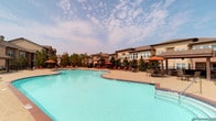 This outdoor pool area offers a generously sized, freeform swimming pool with crystal-clear turquoise water, designed to serve as a central recreational amenity. The pool features a classic brick-tiled border and entry steps, providing easy access from multiple points along its curving perimeter.

Surrounding the pool is a wide, open sundeck finished in a warm aggregate surface, offering ample space for lounging and socializing. The deck is well-furnished with rows of cushioned chaise lounges paired with vibrant orange market umbrellas, providing comfortable shaded retreats throughout the day. Additional seating is available at a dedicated outdoor dining area, equipped with tables and chairs for casual gatherings.

Mature trees and neatly maintained landscaping line the perimeter of the pool area, adding greenery and a sense of privacy. The backdrop features a multi-story residential building with a neutral earth-tone and tan exterior, giving the space a cohesive, community-oriented character. The open-air setting allows for abundant natural light and unobstructed sky views, enhancing the overall resort-like ambiance. The space is well-suited for both relaxed sunbathing and active recreation, making it a functional and inviting outdoor destination.