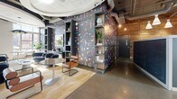 Located at 318 I St NE in Washington, DC, this single-floor amenity space at The Axis offers a thoughtfully designed, open-plan interior that blends contemporary aesthetics with functional versatility. The polished concrete floors reflect abundant natural light streaming through expansive floor-to-ceiling glass walls and doors, which also provide views of an outdoor courtyard area. Exposed ductwork and industrial-style ceilings contrast beautifully with sculptural curved ceiling elements that add architectural interest overhead. A bold, colorful graphic feature wall serves as a striking focal point near the entrance, complemented by built-in shelving displaying curated décor. The space accommodates a variety of uses, with a casual lounge area furnished with rounded blue velvet chairs and bistro-style tables, as well as a more formal conference zone featuring a large oval table surrounded by upholstered seating and a wall-mounted display screen framed by a textured stone accent wall. A reception counter anchors the central area, while pendant lighting and warm wood wall panels add depth and warmth throughout. Lush potted plants are positioned throughout, enhancing the welcoming, vibrant ambiance of this well-appointed community space.