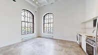 Located on W Lawrence Ave in Chicago's Uptown neighborhood, this single-floor unit is housed within a building of clear architectural character, blending historic detailing with modern interior finishes. The space immediately distinguishes itself through a series of tall, arched windows featuring decorative leaded diamond-pane glazing. These prominent windows flood the interior with natural light and frame views of the surrounding streetscape, creating an open, airy atmosphere throughout.

Polished concrete floors extend consistently across the entire unit, complementing the bright white walls and contributing to the industrial-loft aesthetic. Soaring ceiling heights amplify the sense of volume, particularly in the main living area, where exposed structural elements and track lighting add to the contemporary character of the space.

The kitchen area is efficiently designed, finished with white cabinetry, subway tile backsplash, stainless steel appliances including a refrigerator, range, and dishwasher, and an in-unit washer. The sleeping areas feature generous closet space, with built-in shelving and railing systems. One bedroom benefits directly from an arched leaded window, enhancing its sense of brightness and scale.

The bathroom is cleanly finished in white, equipped with a walk-in shower, vanity with white countertop, mirrored medicine cabinet, and concrete flooring consistent with the rest of the unit.