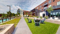 Located at 4066 Lexington Ave N in Shoreview, Minnesota, this generously sized outdoor amenity space at Loma offers a thoughtfully designed environment that blends contemporary architecture with inviting landscaping. The grounds are framed by a modern multi-story building featuring warm wood cladding, white brick masonry, and metal panel accents, creating a striking architectural backdrop throughout the space.

The outdoor area is organized around a central lawn of manicured turf, providing open green space ideal for lawn games such as cornhole. Stamped concrete pathways wind through the grounds, connecting various activity zones seamlessly. A dedicated bocce ball court adds to the recreational offerings, while a cozy fire pit lounge area with Adirondack chairs invites relaxation and social gathering.

Raised concrete and masonry planters are positioned throughout, filled with ornamental grasses, shrubs, and young trees that provide greenery and a sense of enclosure without sacrificing openness. A gated pool area is accessible from the grounds, visible through the perimeter fencing alongside additional landscaped planting beds.

A covered outdoor pavilion with large glass sliding doors connects the interior amenity spaces to the courtyard, enhancing indoor-outdoor flow. The entire space benefits from abundant natural light and open sky views, creating a welcoming community-focused environment year-round.