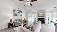 Located at 8755 Jenny Lind St in North Charleston, SC, this single-floor community clubhouse and leasing office offers a well-organized, multifunctional layout designed to welcome residents and visitors alike. Upon entry, a reception area greets guests with a dedicated workstation enclosed by a partitioned office nook, accented by framed artwork and a water cooler. The space transitions seamlessly into a spacious lounge area featuring soaring vaulted ceilings, ceiling fans, and an abundance of natural light streaming through large sliding glass doors that open to views of the outdoor pool. A shiplap-clad fireplace with a brick hearth serves as a striking focal point in the lounge, complemented by plush seating arrangements and tasteful wall art. Neutral carpet flooring runs throughout, contributing to a clean and cohesive aesthetic. A kitchen area with warm wood cabinetry, tile backsplash, and a breakfast bar fitted with vibrant teal bar stools adds a functional and lively touch. A coffee and refreshment station nearby enhances the community-focused atmosphere. Coastal-themed wall décor, exposed beams, and curtained partitions add character, while interior corridors provide access to additional rooms, completing this versatile and inviting community space.
