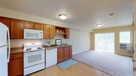This single-floor residential unit offers a thoughtfully designed layout that combines functionality with comfortable living. The kitchen is a standout feature, equipped with warm honey-toned wood cabinetry, ample upper and lower storage, open display shelving, and a full suite of white appliances including a refrigerator, electric range, over-the-range microwave, and dishwasher. Light vinyl flooring runs throughout the kitchen, providing a clean, easy-to-maintain surface. The kitchen opens directly into a generously sized living area, which is finished with plush carpeting and painted in warm neutral tones. A sliding glass door in the living room leads to a private balcony, allowing natural light to fill the space and providing an outdoor view. A wall-mounted climate control unit ensures year-round comfort. The bedroom is carpeted and features a double-door closet with built-in shelving and hanging rods, offering practical storage. The bathroom is designed for convenience, featuring an extended wood vanity with a single-basin sink, a wide mirror with overhead lighting, and a shower/tub combination with light vinyl flooring. The overall ambiance of the unit is clean, neutral, and move-in ready.