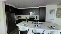 This single-floor space showcases a sleek, contemporary kitchen designed with a striking contrast between dark espresso cabinetry and crisp white countertops. Floor-to-ceiling dark cabinets line the walls, providing generous storage throughout, while recessed lighting illuminates the space from above. The kitchen is equipped with stainless steel appliances, including a gas range, over-the-range microwave, and a full-size refrigerator, offering a fully functional cooking environment. A large, curved white peninsula serves as the focal point of the space, featuring a double undermount sink with a modern faucet, additional prep space, and casual bar seating for two or more, making it ideal for both meal preparation and informal dining. The dark hardwood-style flooring anchors the space and complements the bold cabinetry. The layout is thoughtfully designed in an L-shape, maximizing efficiency and flow. Small decorative plants add subtle warmth and contrast to the otherwise bold, monochromatic palette, contributing to a clean, contemporary ambiance throughout.