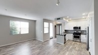 This single-floor space showcases a thoughtfully designed open-concept layout that seamlessly connects the living area and kitchen, creating a bright and functional environment. The kitchen is a standout feature, outfitted with stainless steel appliances including a range and dishwasher, white shaker-style cabinetry, a dark stone countertop, and a classic subway tile backsplash. A central kitchen island with a built-in sink provides additional workspace and serves as a natural dividing point between the cooking and living areas. The living space is finished with warm hardwood-style flooring that runs throughout, contributing to a cohesive and inviting atmosphere. Neutral-toned walls complement the light and airy feel, while multiple windows with blinds allow ample natural light to fill the interior. Recessed and track lighting provide well-distributed illumination overhead. A door visible in the background hints at additional utility or storage space. The overall design balances contemporary aesthetics with practical functionality, making the space feel open, clean, and move-in ready.