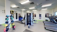 This single-floor fitness facility is designed to support a comprehensive range of training activities. The main workout area is finished with clean white walls and a combination of rubber mat flooring and smooth-surface zones, providing both durability and versatility underfoot. Overhead, multiple ceiling fans and fluorescent lighting fixtures ensure the space remains well-ventilated and brightly illuminated throughout.

The room is outfitted with an impressive variety of equipment. A multi-station strength machine serves as a central fixture, while a heavy punching bag, TRX suspension trainers, and an assortment of medicine balls cater to functional and combat-style training. Foam plyo boxes add options for plyometric and agility work, and additional benches and accessories are positioned throughout the space for flexibility in programming.

A connecting door provides access to an adjacent room, visible through a glass panel, suggesting additional training or rehabilitation space beyond. The overall layout is open and spacious, allowing for comfortable movement between stations. The facility's bright, clean aesthetic and thoughtful equipment arrangement make it well-suited for personal training, group fitness sessions, or physical rehabilitation programs.