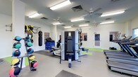 This single-floor fitness facility is designed to support a comprehensive range of training activities. The main workout area is finished with clean white walls and a combination of rubber mat flooring and smooth-surface zones, providing both durability and versatility underfoot. Overhead, multiple ceiling fans and fluorescent lighting fixtures ensure the space remains well-ventilated and brightly illuminated throughout.

The room is outfitted with an impressive variety of equipment. A multi-station strength machine serves as a central fixture, while a heavy punching bag, TRX suspension trainers, and an assortment of medicine balls cater to functional and combat-style training. Foam plyo boxes add options for plyometric and agility work, and additional benches and accessories are positioned throughout the space for flexibility in programming.

A connecting door provides access to an adjacent room, visible through a glass panel, suggesting additional training or rehabilitation space beyond. The overall layout is open and spacious, allowing for comfortable movement between stations. The facility's bright, clean aesthetic and thoughtful equipment arrangement make it well-suited for personal training, group fitness sessions, or physical rehabilitation programs.