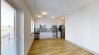 This single-floor space features a clean, contemporary design with an open layout that connects the kitchen and living areas seamlessly. The kitchen is a standout element, equipped with stainless steel appliances including a refrigerator, range with oven, and built-in microwave. Crisp white cabinetry extends from counter height to the ceiling, maximizing storage while contributing to a bright, airy atmosphere. A textured mosaic tile backsplash in muted grey and green tones adds visual interest and contrast against the white surfaces. The countertops provide ample workspace, and a stainless steel sink is positioned beneath a window that allows natural light to filter in. Flush-mount ceiling fixtures provide consistent overhead lighting throughout. Light-toned hardwood flooring runs continuously across the space, enhancing the sense of openness and warmth. The walls are finished in white, keeping the aesthetic minimal and versatile. The overall ambiance is modern and functional, with a neutral palette that offers flexibility for a variety of interior styles.