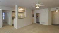 This single-floor residence features an open-concept layout that connects the main living area to the kitchen through a convenient pass-through window, providing visual connectivity while maintaining a defined separation between spaces. The living area is finished with wall-to-wall carpeting in a neutral tone, contributing to a comfortable and understated aesthetic. A ceiling fan with an integrated light fixture is centrally positioned overhead, offering both ambient lighting and air circulation. The walls are painted in a clean, neutral white, lending the space a bright and airy feel.

The kitchen, visible through the pass-through opening, is equipped with white cabinetry, a refrigerator, and overhead lighting that illuminates the workspace effectively. A partial wall with a counter ledge defines the kitchen boundary while maintaining an open connection to the living space.

Toward the rear of the unit, a doorway leads to a dedicated utility area housing a washer and dryer, adding practical convenience. A floor-level vent indicates a forced-air HVAC system, and an electrical panel is also visible nearby. The overall layout is functional and straightforward, designed to maximize usable living space.