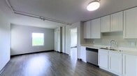 This single-floor unit offers a clean, modern aesthetic with an open-concept design that seamlessly connects the living area and kitchen. The living space is anchored by smooth hardwood-style flooring that runs throughout, complemented by neutral gray walls and crisp white trim that create a light, airy atmosphere. A large window in the living area frames a lush, elevated view of tree canopies, filling the room with abundant natural light. Track lighting runs along the ceiling, providing flexible illumination across the open layout.

The kitchen is efficiently designed with white cabinetry offering ample storage above and below the countertops, a clean light-colored countertop surface, and a stainless steel dishwasher integrated into the lower cabinetry. The kitchen flows naturally into the living space, making it well-suited for both everyday living and entertaining.

A hallway connects the main living area to additional rooms, with multiple white-paneled doors providing access to private spaces within the unit. The overall design is minimalist and functional, with consistent finishes throughout that give the space a cohesive, contemporary feel. The elevated position of the unit, suggested by the treetop views, adds a sense of openness and tranquility to the interior ambiance.