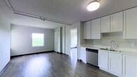 This single-floor unit offers a clean, modern aesthetic with an open-concept design that seamlessly connects the living area and kitchen. The living space is anchored by smooth hardwood-style flooring that runs throughout, complemented by neutral gray walls and crisp white trim that create a light, airy atmosphere. A large window in the living area frames a lush, elevated view of tree canopies, filling the room with abundant natural light. Track lighting runs along the ceiling, providing flexible illumination across the open layout.

The kitchen is efficiently designed with white cabinetry offering ample storage above and below the countertops, a clean light-colored countertop surface, and a stainless steel dishwasher integrated into the lower cabinetry. The kitchen flows naturally into the living space, making it well-suited for both everyday living and entertaining.

A hallway connects the main living area to additional rooms, with multiple white-paneled doors providing access to private spaces within the unit. The overall design is minimalist and functional, with consistent finishes throughout that give the space a cohesive, contemporary feel. The elevated position of the unit, suggested by the treetop views, adds a sense of openness and tranquility to the interior ambiance.