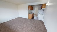 Located at 8101 SW 83rd Ave, Unit D22 in Lakewood, Washington, this single-floor residence offers a straightforward and functional layout designed for comfortable everyday living. The living area features neutral wall-to-wall carpeting that creates a warm and inviting atmosphere, providing a versatile space suitable for a variety of furniture arrangements. The open sightline from the living area into the kitchen enhances the sense of space and connectivity throughout the unit. The kitchen is equipped with warm-toned wooden cabinetry both above and below the counters, offering ample storage for daily needs. A double-basin sink is centrally positioned beneath a window that allows natural light to brighten the workspace. The kitchen also features a white electric range and a full-size white refrigerator, providing all the essential appliances for meal preparation. The kitchen flooring transitions to a dark hardwood-style surface, creating a clear and practical distinction from the carpeted living area. A textured ceiling is consistent throughout the unit, adding a subtle architectural detail. The layout is efficiently organized across a single level, making it easily accessible and simple to navigate.