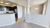This single-floor space showcases an open-concept design that connects the living area and kitchen in a cohesive, functional layout. A prominent decorative column with a classical base and capital serves as a striking architectural focal point, adding a touch of elegance to the interior while subtly defining the transition between spaces.

The living area features neutral-toned carpeting that lends a warm, comfortable feel underfoot. Soft, neutral wall colors throughout create a clean and inviting atmosphere, complemented by crisp white baseboards and trim. A chandelier with multiple candle-style lights adds a refined touch to the dining or transitional area, enhancing the overall aesthetic.

The kitchen is visible from the living space and features dark cabinetry that contrasts sharply against light countertops, creating a bold, modern look. Stainless steel appliances, including a refrigerator, contribute to the kitchen's contemporary character. Wood-style flooring in the kitchen distinguishes it from the carpeted living area, providing a practical and visually appealing transition between the two zones. Overall, the space presents a balanced combination of classic architectural details and modern finishes.