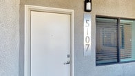 This single-story space is accessed through a clean white entry door set against a textured stucco exterior, with a sleek vertical metal address plate and contemporary wall-mounted light fixture marking the entrance. The exterior presents a neat, modern curb appeal with crisp lines and a well-maintained facade.

Inside, the open-concept layout connects the living, dining, and kitchen areas in one cohesive, light-filled space. The kitchen serves as a focal point, featuring dark espresso cabinetry that contrasts sharply with a bright white island with a waterfall-style surround. Stainless steel appliances, including a French door refrigerator and range, are complemented by a wall-mounted range hood and stylish pendant lighting suspended above the island. Recessed lighting throughout the ceiling provides even, modern illumination.

The living area is anchored by a ceiling fan with integrated lighting, and light-toned wood-look flooring runs throughout, lending warmth and visual continuity. Black-framed interior doors add a bold, contemporary accent. A dining area with natural window light sits adjacent, completing the open-plan arrangement. The overall ambiance is modern, clean, and well-appointed.