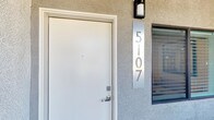 This single-story space is accessed through a clean white entry door set against a textured stucco exterior, with a sleek vertical metal address plate and contemporary wall-mounted light fixture marking the entrance. The exterior presents a neat, modern curb appeal with crisp lines and a well-maintained facade.

Inside, the open-concept layout connects the living, dining, and kitchen areas in one cohesive, light-filled space. The kitchen serves as a focal point, featuring dark espresso cabinetry that contrasts sharply with a bright white island with a waterfall-style surround. Stainless steel appliances, including a French door refrigerator and range, are complemented by a wall-mounted range hood and stylish pendant lighting suspended above the island. Recessed lighting throughout the ceiling provides even, modern illumination.

The living area is anchored by a ceiling fan with integrated lighting, and light-toned wood-look flooring runs throughout, lending warmth and visual continuity. Black-framed interior doors add a bold, contemporary accent. A dining area with natural window light sits adjacent, completing the open-plan arrangement. The overall ambiance is modern, clean, and well-appointed.