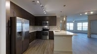 This single-floor residence showcases a thoughtfully designed open-concept layout that blends style and functionality. The kitchen is a standout feature, outfitted with sleek dark cabinetry that contrasts beautifully against crisp white quartz countertops and a classic white tile backsplash. Stainless steel appliances — including a French door refrigerator, electric range, and built-in microwave — lend a contemporary, professional feel to the space. A large central island with an undermount sink serves as both a practical workspace and a natural gathering point, illuminated by elegant pendant lighting that hangs from above. Track lighting along the ceiling provides additional brightness throughout the kitchen. The warm-toned luxury vinyl plank flooring runs continuously from the kitchen into the adjacent living area, reinforcing the seamless open-concept flow. Natural light pours in through windows in the living space, enhancing the airy and inviting atmosphere. A ceiling fan in the living area adds to the comfort of the home. The overall design aesthetic is modern and cohesive, combining dark and light tones to create a balanced, welcoming environment ideal for everyday living and entertaining.
