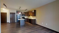 Located at 310 Samuel Dr in Madison, Wisconsin, this single-floor residence offers a thoughtfully designed open-concept layout that seamlessly blends living and kitchen spaces. The kitchen is a standout feature, showcasing rich dark wood cabinetry that extends to the upper walls, providing generous storage throughout. Light-toned countertops offer a striking contrast against the darker cabinetry, and a central island serves as both a prep space and a casual gathering point. Pendant lighting above the island adds a warm, focused glow, complemented by a track lighting system overhead that illuminates the full kitchen area. Black appliances, including a refrigerator, range, and dishwasher, maintain a cohesive and modern aesthetic. The sink is positioned along a dedicated counter run, offering a practical workspace with additional lower cabinetry. Dark hardwood-style flooring spans the entire visible area, lending warmth and continuity to the space. The open layout allows natural sightlines from the kitchen into the adjoining living area, creating an airy and connected atmosphere. Neutral wall tones in warm beige enhance the brightness of the space and provide a versatile backdrop. Overall, the unit combines functional design with modern finishes for comfortable everyday living.