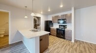 This single-floor residence offers a thoughtfully organized layout comprising two bedrooms, two bathrooms, a kitchen, a living room, a dedicated laundry area, and a closet. The open-concept kitchen serves as a central hub, featuring a peninsula island with a double sink and light countertops, rich dark-stained cabinetry, black appliances including a refrigerator and range, and pendant lighting overhead. Recessed lighting and wood-look luxury vinyl flooring carry throughout the kitchen and common areas, contributing to a clean, cohesive aesthetic.

The living room is carpeted and benefits from a sliding glass door that invites natural light and provides access to an outdoor area with neighborhood views. Both bedrooms are carpeted and finished with neutral tones; the secondary bedroom includes a window with exterior views, while the primary bedroom features a dedicated closet with built-in wire shelving. The two bathrooms are finished with wood-look vinyl flooring, dark vanity cabinetry, and full tub-shower combos with curved curtain rods. The laundry area houses the in-unit furnace and accommodates a washer and dryer. Overall, the space presents a functional and practical living environment with consistent, contemporary finishes.