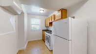 This single-floor space presents a clean, straightforward layout spread across several interconnected rooms. A defining characteristic throughout the unit is the warm-toned parquet wood flooring, which adds a classic touch to each room and reflects natural light to brighten the interiors.

The living area is a well-proportioned space featuring a wall-mounted HVAC unit, a ventilation grille, and a window fitted with blinds that allows greenery views from outside. Adjacent to the main living area, the kitchen is neatly arranged with honey-toned upper cabinetry, a dark granite-style countertop, a gas range, and a full-size refrigerator. Tile flooring and a window overlooking outdoor greenery complete the kitchen's functional design.

The remaining rooms share a consistent aesthetic: white walls, parquet flooring, and individual windows that invite natural light and outdoor views. One room benefits from dual windows positioned on adjacent walls, significantly brightening the space. Each room is accessed via standard interior doors, and the overall flow between spaces is straightforward and practical. Ceiling-mounted light fixtures are present throughout, ensuring consistent illumination across the unit.