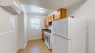 This single-floor space presents a clean, straightforward layout spread across several interconnected rooms. A defining characteristic throughout the unit is the warm-toned parquet wood flooring, which adds a classic touch to each room and reflects natural light to brighten the interiors.

The living area is a well-proportioned space featuring a wall-mounted HVAC unit, a ventilation grille, and a window fitted with blinds that allows greenery views from outside. Adjacent to the main living area, the kitchen is neatly arranged with honey-toned upper cabinetry, a dark granite-style countertop, a gas range, and a full-size refrigerator. Tile flooring and a window overlooking outdoor greenery complete the kitchen's functional design.

The remaining rooms share a consistent aesthetic: white walls, parquet flooring, and individual windows that invite natural light and outdoor views. One room benefits from dual windows positioned on adjacent walls, significantly brightening the space. Each room is accessed via standard interior doors, and the overall flow between spaces is straightforward and practical. Ceiling-mounted light fixtures are present throughout, ensuring consistent illumination across the unit.