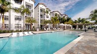 This outdoor amenity space is defined by its Mediterranean-inspired architectural aesthetic, showcased through crisp white stucco building facades, terracotta-tiled rooflines, and elegant arched detailing. At the heart of the space lies a generously sized swimming pool with crystal-clear blue water, surrounded by a broad paver deck finished in warm, multi-toned stone. Rows of white lounge chairs line the poolside, offering ample space for sunbathing, while multiple white fabric shade canopies and umbrellas provide sheltered relaxation areas throughout the deck.

Tall, mature palm trees are strategically placed across the grounds, lending a tropical, resort-like atmosphere and natural shade. Well-maintained landscaping with lush greenery and decorative stone accents borders the pool area, enclosed by a sleek black wrought-iron fence that maintains both security and an open, airy feel.

A covered pavilion area features distinctive arched entryways, a warm wood-paneled ceiling, ceiling fans, and paver flooring, offering an elegant transitional space between indoors and the pool deck. Oversized glass doors and windows connect interior spaces to the outdoors seamlessly. Open views beyond the perimeter reveal tall palms against a bright Florida sky, reinforcing the relaxed, upscale ambiance of this well-appointed outdoor retreat.