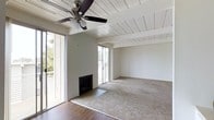 This single-floor space offers a bright and open living area defined by its distinctive white-painted wood-planked ceiling with exposed beams, lending a warm, mid-century character to the interior. A ceiling fan is centrally mounted, providing both functional airflow and a classic aesthetic touch. The living area is anchored by a fireplace set against the wall, with a tiled hearth that transitions into rich hardwood flooring near the entry zones, while plush carpeting covers the main living space for added comfort. Two sets of sliding glass doors are positioned on adjacent walls, flooding the room with generous natural light and offering views of the surrounding greenery outside. The overall palette is kept neutral and clean, with crisp white walls that complement the light-toned ceiling and allow for versatile interior styling. The open layout and thoughtful architectural detailing create a welcoming ambiance that balances casual comfort with understated elegance, making the space feel airy and expansive throughout.