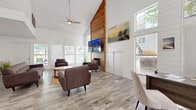 This single-floor open-concept space combines a living room and kitchen into a cohesive, well-designed layout. The living area immediately draws the eye upward with its dramatic vaulted ceilings, clad in crisp white shiplap that enhances the bright, airy atmosphere. A standout architectural feature is the floor-to-ceiling natural wood accent wall, which frames a wall-mounted flat-screen TV and a classic fireplace below, creating a warm and stylish focal point. Abundant natural light pours in through multiple large windows fitted with Venetian blinds, offering views of the surrounding greenery. The space is finished with light wood-tone luxury vinyl plank flooring throughout, and a ceiling fan adds both function and a relaxed coastal charm. The seating arrangement features mid-century modern upholstered sofas and armchairs centered around a round wooden coffee table, complemented by a woven area rug. The kitchen is designed with clean white shaker cabinetry, a white subway tile backsplash, a large island with a grey quartz countertop and integrated sink, and a full suite of stainless steel appliances. Track lighting above the kitchen ensures excellent task illumination. Together, the two spaces deliver a cohesive coastal-modern aesthetic that is both functional and inviting.