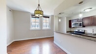 Located at 617 W Lexington St in Baltimore, MD, this single-floor residence offers a thoughtfully designed layout that balances functionality and style. The open-concept living and dining area is anchored by rich hardwood flooring and framed by two large windows that allow natural light to pour in, offering views of the surrounding urban streetscape. A decorative pendant light fixture adds a stylish focal point to the dining area. The kitchen is separated from the living space by a half-wall peninsula with granite countertops, creating an efficient and connected cooking environment. Dark espresso cabinetry pairs with stainless steel appliances — including a range, microwave, dishwasher, and refrigerator — to deliver a modern, functional culinary space. The bedroom features plush wall-to-wall carpeting, a window overlooking the neighborhood, and a closet with built-in shelving. A dedicated in-unit laundry area with stacked washer and dryer adds everyday convenience. The bathroom is finished with warm travertine-style tile throughout, complemented by a glass-enclosed walk-in shower, dark wood vanity cabinetry, and clean chrome fixtures. A fireplace visible in the dollhouse view adds a welcoming architectural touch to the living area, completing this urban Baltimore residence.