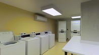 This shared laundry facility is designed with functionality and convenience in mind. The room is finished with warm yellow walls and a light-toned floor, creating a clean and welcoming environment. Multiple top-loading washing machines are arranged in a row along one wall, providing ample capacity for residents or guests. Stacked dryer units are positioned toward the back of the space, maximizing the use of vertical space and allowing for efficient organization of equipment. A large, white folding table is positioned to one side, offering a dedicated surface for sorting and folding laundry. Overhead, a large flat-panel LED light fixture provides bright, even illumination throughout the room, supplemented by additional lighting toward the rear. A wall-mounted split-system air conditioning unit ensures the space remains comfortable during use. The layout is practical and open, allowing for easy movement between machines. Overall, the facility is well-maintained and thoughtfully arranged to serve the needs of multiple users efficiently.