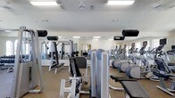This fitness center offers a thorough and well-organized workout facility designed to accommodate a wide range of exercise needs. The space is outfitted with a robust selection of strength-training equipment, including multi-function cable machines, adjustable weight benches, and resistance machines targeting various muscle groups. On the cardio side, a row of modern elliptical trainers lines one section of the room, providing ample options for aerobic exercise.

The interior features bright, recessed fluorescent lighting that evenly illuminates the entire floor, creating a clean and energizing atmosphere. Full-length mirrors span the walls, visually expanding the space while allowing users to monitor their form during workouts. The neutral-toned flooring complements the light-colored walls and white ceiling, contributing to a tidy, professional environment.

The layout is thoughtfully arranged, with strength equipment and cardio machines organized into distinct zones to promote efficient use of the space and easy navigation between stations. The overall ambiance is functional and focused, making it well-suited for both casual exercisers and more dedicated fitness enthusiasts.