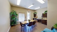 This single-floor community space is thoughtfully designed to serve as a functional and welcoming gathering area. The main room is anchored by a natural wood dining table surrounded by dark leather chairs, providing an inviting setting for meetings or shared meals. Wood-look tile flooring runs throughout, adding warmth and a cohesive aesthetic to the space.

Along one wall, a well-equipped kitchenette features rich, dark espresso cabinetry paired with light granite countertops, an undermount sink, and ample upper and lower cabinet storage. Lush green plants are displayed atop the cabinetry, adding a fresh, vibrant touch to the room.

A lounge area offers teal upholstered seating alongside a colorful piece of wall art, creating a relaxed corner for informal conversations. Recessed and overhead fluorescent lighting ensures the space is brightly and evenly lit throughout.

Natural light enters through a large window fitted with horizontal blinds, and a glass-panel exterior door opens to an outdoor area, offering views of a sunny courtyard or common grounds beyond. The neutral beige wall tones throughout keep the atmosphere calm and versatile, making this a practical and pleasant space for a variety of communal activities.