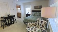 This single-floor unit offers an open, functional layout that combines the sleeping and living areas into one cohesive space. The bedroom area is carpeted and comfortably accommodates a full-sized bed with a dark wood headboard, complemented by a matching nightstand with a table lamp. A patterned accent chair adds a decorative touch to the room, while a small desk or bar-height table near the wall provides a practical workspace or dining spot. A window with a roller shade allows natural light to brighten the space. Wall-mounted artwork above the bed adds a finishing detail to the room. Directly accessible through an open doorway, the kitchen features wooden cabinetry, a countertop with a sink, and what appears to be standard appliances. The transition between the two spaces is seamless, contributing to the unit's open and connected feel. The neutral wall tones and light carpet throughout create a clean, cohesive aesthetic. Overall, the space is thoughtfully arranged to maximize comfort and utility within a compact footprint.