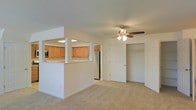 This single-floor space features a practical, open-concept layout that connects the living area and kitchen seamlessly. The living area is finished with neutral wall-to-wall carpeting, contributing to a clean and comfortable atmosphere. A ceiling fan with an integrated light fixture is centrally positioned, providing both air circulation and overhead lighting throughout the space. A spacious walk-in closet with a full-panel door offers generous built-in storage, with a closet rod and shelving visible inside.

The kitchen is accessible via a pass-through counter with a breakfast bar-style ledge, framed by a partial wall that defines the space without fully enclosing it. Inside the kitchen, warm honey-toned cabinetry lines the upper walls, paired with light countertops and a stainless steel refrigerator. Recessed lighting brightens the kitchen area, enhancing the visibility and functionality of the workspace. The overall design emphasizes efficiency and ease of use, with a neutral color palette of whites and beiges throughout that gives the space a bright, airy feel well-suited for everyday living.