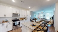 This single-floor residence showcases a thoughtfully designed open-concept layout that seamlessly connects the kitchen, dining, and living areas. The living room is defined by a signature teal accent wall, wood-look flooring, and abundant natural light streaming through large windows dressed with blinds and warm-toned drape panels. A ceiling fan with an integrated light fixture anchors the space, while gold-toned sconce lighting adds a refined touch. A compact dining area with a round table sits adjacent to the front entry door, making efficient use of the layout.

The kitchen is well-equipped with white shaker-style cabinetry, granite countertops, stainless steel appliances including a range, microwave, and refrigerator, and a central island with a built-in sink and bar seating. Recessed lighting illuminates the work surfaces throughout.

The bedroom offers a ceiling fan, wood-look flooring, and a large window with outdoor views, creating a calm and restful atmosphere. The bathroom features a granite-topped vanity, undermount sink, and wood-look flooring, maintaining the consistent finish quality found throughout the residence.