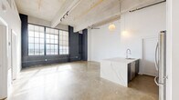 Located at 2000 North 2nd Street in Philadelphia, PA, this 1-bedroom, 2-bathroom loft seamlessly blends industrial heritage with contemporary design. The open-plan living area commands attention with soaring ceilings, exposed concrete beams, and original painted brick columns, all bathed in abundant natural light pouring through expansive factory-style, floor-to-ceiling windows that frame sweeping views of the Philadelphia skyline. Polished concrete floors flow throughout, lending a cohesive and modern industrial aesthetic to every room. The kitchen serves as a design centerpiece, anchored by a large waterfall-edge quartz island with an undermount sink and a professional-style pull-down faucet. Sleek two-tone cabinetry — white uppers paired with dark lowers — complements a full suite of stainless steel appliances, including a French door refrigerator, electric range, and built-in microwave, all set against a classic white tile backsplash. The generously sized bedroom benefits from the same dramatic window presence, with a ceiling fan and ample closet space. Two well-appointed bathrooms continue the clean, minimalist palette, one featuring a glass-enclosed walk-in shower and the other a full soaking tub, both with floating vanities and polished concrete floors.