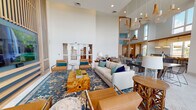 This single-level waterfront space is defined by its dramatic double-height ceilings and an expansive wall of floor-to-ceiling windows that frame sweeping views of the water and a distant bridge beyond. The open-plan layout flows seamlessly between several distinct zones, each thoughtfully furnished to encourage both social interaction and relaxed retreat.

The main lounge area centers on a plush neutral sofa accented with jewel-toned throw pillows, arranged around a striking burl wood coffee table atop a richly patterned blue area rug. A full-height slatted wood and teal accent wall serves as a bold focal point, anchoring a large wall-mounted television flanked by cognac leather armchairs.

Adjacent to the lounge, a long communal dining table with wire-back chairs seats a generous gathering, illuminated by woven pendant lights that echo the coastal aesthetic throughout. A sleek counter-height bar runs along the waterfront windows, lined with woven rattan barstools beneath warm drum pendants with a golden-toned ceiling detail above.

Artful room dividers constructed of wood-framed rope screens create a sense of definition without sacrificing openness. Light washes across whitewashed hardwood floors, enhancing the airy, resort-inspired ambiance that permeates the entire space.