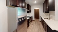 Located at 2050 Delaware Ave in West St. Paul, Minnesota, this single-floor unit offers a practical and comfortable layout with a clean, neutral aesthetic throughout. The living area is generously sized, featuring a combination of plush carpeting and hardwood-style flooring, with a wall-mounted air conditioning unit and access to natural light through a large window that frames views of surrounding greenery. The space transitions seamlessly into a peninsula-style kitchen, which is equipped with dark espresso cabinetry, stainless steel appliances including a refrigerator and range, a wall-mounted range hood, recessed lighting, and light countertops that contrast elegantly with the rich cabinetry tones. The kitchen also features hardwood-style flooring and provides a functional, open connection to the main living space. The bedroom is a quiet, carpeted retreat with a generous closet featuring built-in shelving and a hanging rod, as well as a window with verdant outdoor views and a baseboard heating unit. The full bathroom is finished with white subway tile surrounding the tub-shower combination, a clean toilet, and a dark wood wall-mounted cabinet for storage. Throughout the unit, dark wood trim and baseboards provide a consistent, grounded design detail.