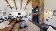 This single-floor amenity space masterfully combines rustic and contemporary design elements to create a welcoming, multi-functional environment. The great room serves as the centerpiece, boasting soaring vaulted ceilings adorned with exposed wood beams, a dramatic floor-to-ceiling stacked stone fireplace, and large windows that flood the space with natural light and offer views of the surrounding greenery. A mounted flat-screen TV and comfortable sectional seating make this area ideal for gatherings and relaxation.

Adjacent to the great room is a game and recreation zone, complete with a pool table, shuffleboard, and a wall-length counter bar with black bar stools positioned in front of expansive windows — providing both entertainment and scenic outdoor views.

The dining area accommodates a large group around a substantial dark wood farmhouse table beneath additional exposed beam detailing and recessed lighting. A wall-mounted TV anchors the space, adding a functional touch.

The fully equipped kitchen features gray and white cabinetry, white subway tile backsplash, open shelving, stainless steel appliances, and a generous island with bar seating — designed for both casual use and catered events. Light hardwood flooring flows consistently throughout, tying the entire space together with a clean, cohesive aesthetic.