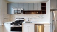 Unit 57 at 518 Orange St in New Haven, Connecticut is a well-appointed 2-bedroom, 2-bathroom residence situated on a single floor. The open layout connects the living area directly to the kitchen, which features contemporary grey gloss upper cabinetry, warm wood-toned open shelving, white lower cabinets, white quartz countertops, a stone tile backsplash, and a full suite of stainless steel appliances including a gas range, built-in microwave, dishwasher, and refrigerator. Rich, dark-stained hardwood floors run throughout the living spaces and both bedrooms, providing a warm contrast to the bright white walls and crisp trim work. Each bedroom is well-sized with natural light from framed windows; one bedroom offers a treetop view of the surrounding New Haven neighborhood. The two bathrooms reflect modern design sensibilities: one includes a floating walnut vanity with a matte black faucet, large-format wall tiles, and a wide mirror, while the second features an open walk-in shower with mosaic tile flooring and contrasting marble-style wall tiles. Classic architectural details such as substantial baseboards and window casings complement the updated interior throughout.