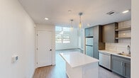 This contemporary two-story residence offers a thoughtfully designed layout that maximizes functionality and style. The main floor serves as the heart of the home, anchored by a sleek, modern kitchen outfitted with gray shaker cabinetry, quartz countertops, a large center island, subway tile backsplash, and a full suite of stainless steel appliances including a gas range, built-in microwave, and dishwasher. Elegant pendant lighting illuminates the island, adding a refined touch to the space. The kitchen flows seamlessly into the open living area, which is bathed in natural light through a striking bank of floor-to-ceiling and clerestory windows that frame outdoor views and create an airy, sun-filled atmosphere. Light wood-look flooring runs throughout both levels, lending warmth and continuity. Upstairs, the private bedroom is a comfortable retreat featuring a ceiling fan, a window with natural light, and neutral toned walls. An adjacent walk-in closet provides ample storage with built-in wire shelving. The full bathroom is finished with clean, floor-to-ceiling white tile surrounding a soaking tub and shower combination with chrome fixtures. A dedicated hallway and staircase connect both levels efficiently, completing this well-appointed modern home.