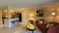 This single-floor residence offers a comfortable and well-appointed layout designed for easy everyday living. The open-concept living area is bright and welcoming, featuring light tile flooring, warm neutral walls, and a textured ceiling that adds subtle character to the space. The living room is anchored by a cozy sofa, an oval glass-top coffee table, and a decorative side lamp, creating an inviting atmosphere ideal for relaxing or entertaining.

A tasteful piece of wall art adds a pop of color and personality to the living space, complementing the warm tones throughout. The layout flows naturally into the kitchen and dining area, which is separated by a breakfast bar with counter-height seating — perfect for casual meals or morning coffee. The kitchen features dark cabinetry, dark appliances, and a functional counter layout that makes efficient use of the available space.

The front entry door is centrally positioned, offering a clean sightline from the main living area. Natural and ambient lighting work together to create a warm glow throughout the space. Overall, the residence presents a cozy, functional, and neatly arranged interior well-suited for comfortable single-floor living.