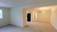 This single-story residence is designed with an open-concept layout that promotes a seamless flow between its main living spaces. The interior is finished in soft, neutral tones—light greens and warm beiges—that lend a calm, airy atmosphere throughout. Plush wall-to-wall carpeting in a light tan hue covers the floors, adding warmth underfoot across the living and dining areas.

A wide archway connects the main living space to the kitchen area, which is visible from across the room. The kitchen features white upper cabinetry and is equipped with a full-size black refrigerator, providing a functional contrast to the lighter surroundings. Adjacent to the kitchen, a hallway leads toward additional rooms, suggesting a practical and efficient floor plan.

The dining area is defined by a classic pendant light fixture that hangs from the ceiling, providing both ambient illumination and a subtle decorative focal point. A window in this area allows natural light to filter into the space. Recessed flush-mount ceiling lighting supplements the overall brightness. White baseboards and clean wall lines throughout contribute to the tidy, well-maintained appearance of this single-level home.