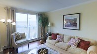 This single-floor space centers around a welcoming living room designed for both comfort and functionality. The room is bathed in natural light streaming through a large sliding glass door that opens onto a private balcony, offering outdoor views and enhancing the sense of openness. Cream-toned walls complement the warm hardwood flooring underfoot, creating a cohesive and inviting atmosphere throughout.

The living area is generously proportioned, accommodating a plush, neutral-toned sofa dressed with decorative red and patterned accent pillows, alongside a cushioned armchair. A soft, textured area rug anchors the seating arrangement, adding warmth and depth to the space. A decorative bowl centerpiece rests on the coffee table, contributing to the curated aesthetic.

Crown molding runs along the ceiling, adding a refined architectural detail to the room. A framed landscape artwork adorns one wall, while a tall indoor plant near the balcony door introduces a natural, organic element. A classic table lamp and a dark wood console table provide both ambient lighting and practical storage, rounding out the well-appointed living space.
