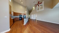 Located at 251 Town Center Ln in Keller, TX, this two-story residence offers a thoughtfully designed layout with a striking open-concept main level. The living and dining area immediately impresses with soaring vaulted ceilings, rich hardwood flooring, and track lighting, while a ceiling fan adds both function and visual interest. A second-floor overlook with a railing is visible from below, reinforcing the dramatic verticality of the space.

The kitchen is well-appointed with warm wood cabinetry extending to the upper reaches of the tall walls, granite countertops, a large central island with a polished stone surface, and black appliances including a range, microwave, dishwasher, and refrigerator. A window above the sink brings in natural light.

A wide hallway on the main level leads to additional rooms, with generous closet space along the way. One bathroom features wood-toned cabinetry, a white countertop, and a tiled tub-shower combination. A second half-bath offers a similar vanity style with hardwood flooring.

Upstairs, carpeted bedrooms provide a comfortable retreat. One room features elegant mirrored French doors, while another offers a ceiling fan and a window with outdoor views. Throughout, a neutral warm-gray palette and consistent trim work create a cohesive, polished aesthetic.