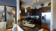 This single-floor space showcases a thoughtfully designed interior with a strong emphasis on modern aesthetics and quality finishes. The kitchen serves as a standout centerpiece, outfitted with rich, dark espresso cabinetry that extends to the ceiling, providing generous storage while creating a sleek, sophisticated look. The countertops are finished in deep granite, offering both durability and visual depth that complements the dark wood tones throughout.

A suite of stainless steel appliances — including a range, built-in microwave, refrigerator with ice dispenser, and additional counter appliances — equips the kitchen for full functionality. A kitchen island with a wraparound granite surface provides additional workspace and casual seating, anchored by stylish studded upholstered seating.

Overhead, a combination of track lighting and striking elongated pendant lights illuminate the space with a warm, dramatic glow. Decorative accents including tall glass vessels, potted greenery atop the cabinetry, and artful dishware add personality and warmth to the kitchen area. An ornamental mirrored panel with an artistic motif near the entry further enhances the space's refined character. The overall ambiance is modern, polished, and inviting.