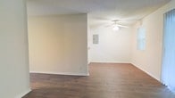 This single-story space offers a straightforward, functional layout highlighted by rich, dark hardwood-style flooring that extends throughout the open living area. The warm tones of the flooring contrast cleanly against the neutral, light-colored walls, creating a balanced and inviting atmosphere. A textured ceiling adds subtle depth overhead, while a ceiling fan with an integrated light fixture provides both practical airflow and ambient lighting. The open floor plan is defined by a partial interior wall that gently divides the space into distinct zones without fully closing off the layout, allowing for flexible furniture arrangement and a sense of continuity. White baseboards frame the perimeter of the room, adding a crisp, clean finish to the overall aesthetic. An electrical panel is visibly mounted on the wall, a practical feature that speaks to the functional nature of the space. The interior is well-maintained, freshly painted, and move-in ready, making it a versatile option suited to a variety of uses. Overall, the space presents a clean, simple design that prioritizes usability and ease of personalization.