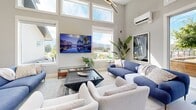 This thoughtfully designed single-floor amenity space offers a bright and welcoming atmosphere, highlighted by dramatic double-height ceilings and an abundance of natural light streaming through expansive clerestory and large picture windows. The open-concept layout seamlessly connects the living and recreational zones, which feature comfortable lounge seating, a wall-mounted flat-screen TV, curated artwork, and a full-size pool table — creating an inviting setting for both relaxation and socializing. A modern kitchen anchors one side of the space, outfitted with sleek, high-gloss white cabinetry, stainless steel appliances including a range, dishwasher, and refrigerator, and a generous dining table with leather-accented chairs. Light wood-tone flooring flows throughout, complementing the neutral grey walls and clean contemporary aesthetic. A wall-mounted mini-split system provides climate control, while a sculptural multi-arm pendant light adds an artistic focal point overhead. The space also includes a fully accessible washroom equipped with grab bars and compliant fixtures. Views of surrounding greenery and distant mountains can be seen through the many windows, enhancing the airy, light-filled ambiance of this well-appointed communal space.
