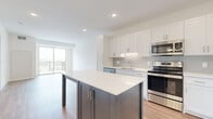 Located at 10010 6th Ave N in Plymouth, Minnesota, this thoughtfully designed single-floor unit offers a clean, modern aesthetic with high-quality finishes throughout. The open-concept kitchen serves as the heart of the space, featuring crisp white shaker-style cabinetry, a subtle tile backsplash, gleaming white quartz countertops, and a full suite of stainless steel appliances including a range, microwave, dishwasher, and refrigerator. A contrasting dark-framed center island provides additional prep and dining space, adding visual interest to the layout. The kitchen flows seamlessly into a bright living area bathed in natural light from large sliding glass doors that open onto a private balcony, offering outdoor views of the surrounding Plymouth area. Durable light-toned hardwood-style flooring runs throughout the main living spaces, while the bedroom features plush neutral carpeting and a generous window that invites abundant natural light. The bathroom is well-appointed with white shaker vanity cabinetry, a dual-sink white countertop, a large mirror, and warm wood-tone flooring. The unit's clean white walls, recessed lighting, and consistent modern hardware create a cohesive, contemporary ambiance throughout.