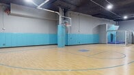 This indoor futsal facility offers a generously sized, single-level playing surface covered in green artificial turf, complete with clearly marked white boundary lines, a center circle, and goal boxes at each end. Two full-size portable goals are positioned at opposing ends of the pitch, making the space ready for competitive or casual play. The structural aesthetic is defined by exposed concrete walls and ceiling, lending an industrial character that is both raw and functional. Overhead, a grid of recessed and surface-mounted fluorescent panel lights ensures even, bright illumination across the entire playing area. One wall is dramatically transformed by a large-scale graphic mural featuring bold blue and white geometric shapes — hexagons and sharp angular forms — alongside the energetic phrase "Live, Work, Play" rendered in dynamic lettering. This eye-catching artwork adds a vibrant, motivational atmosphere to the space. Along one side wall, player benches and a utility rack provide practical amenities for teams and participants. The space is well-suited for organized matches, training sessions, or recreational leagues, combining athletic functionality with a visually engaging environment that encourages active use.